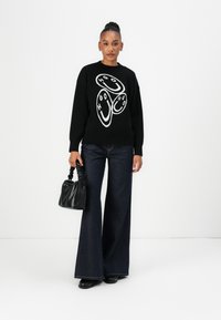 Zwarte gebreide trui met witte grafische ontwerp aan de voorkant, gecombineerd met donkerblauwe hoge taille flared jeans. Accessoire: kleine zwarte handtas.
