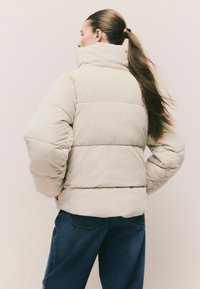 Beige Cordhose Pufferjacke mit hohem Kragen, ausgestattet mit einem gesteppten Design und puffigen Ärmeln. Getragen mit dunkelblauen weiten Jeans.