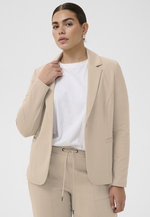 Woman wearing beige blazer and matching pants with white drawstring, white t-shirt, holding blazer lapel against plain background.