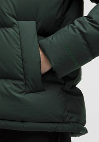 Tmavo zelená prešívaná bunda s rukou vo prednej vrecku a brandingom "Lululemon" na manžete rukávu.