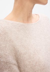 Gros plan d'une personne portant un pull en tricot beige clair avec un large col rond et une zone visible du cou et des épaules.