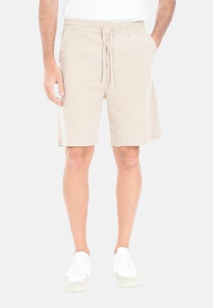 Uomo che indossa pantaloncini beige con coulisse e tasche, e sneaker bianche, in piedi contro uno sfondo chiaro e semplice.