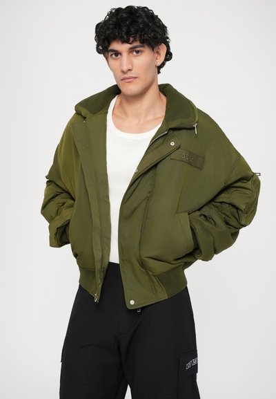 Khaki bomberjack met een ritssluiting en drukknoopsluiting. Heeft oversized mouwen en een contrasterende groene kraag. Gedragen over een wit overhemd.
