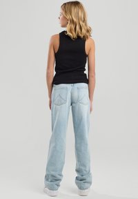 Young woman with blonde hair wearing a black sleeveless top, light blue loose jeans, and white sneakers, standing facing backward on gray background.