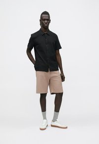 Zwart gestreept T-shirt met korte mouwen, beige korte broek, grijze sokken, witte sneakers met bruine zolen. Model staat tegen een effen witte achtergrond.