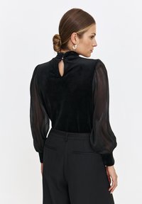 Czarny top z fakturowanym welurowym korpusem i przezroczystymi bufkami. Posiada dekolt w kształcie łezki z małym zapięciem na guzik. W połączeniu z czarnymi spodniami.