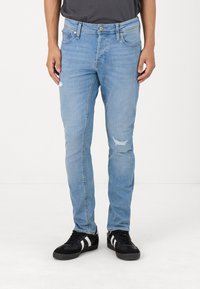 Helle blaue Denim-Jeans im Slim Fit mit distressed Details an den Knien und einem Fünf-Taschen-Design. Getragen mit schwarzen Schuhen.