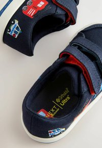 Marina canvas skor med kardborreband, prydda med broderade brandbil- och polisbilsmönster. Vit gummisula med blå detaljer.