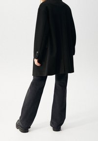 Cappotto oversize in misto lana nero con colletto a punta e chiusura a tre bottoni, abbinato a jeans neri a gamba larga e stivali neri chunky.