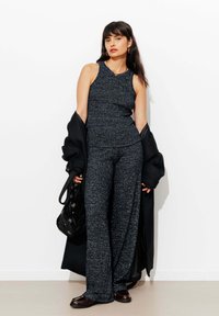 Ensemble en deux pièces composé d'un haut ajusté sans manches de couleur bleu foncé et d'un pantalon large, texturé avec des accents de fil métallique subtils. Un manteau noir tombe sur un bras.