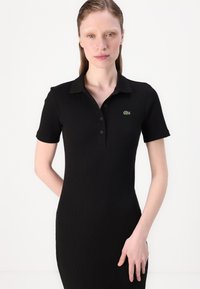 Juoda raštuota polo suknelė su trumpomis rankovėmis, apykakle, sagų sąsaja ir mažas žalias logotipas kairėje krūtinės pusėje. Lygus paviršius.