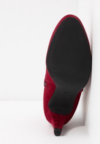 Chaussure en daim rouge à bout pointu inclinée vers le haut, montrant la semelle noire texturée avec un petit talon sur un fond blanc d'angle.