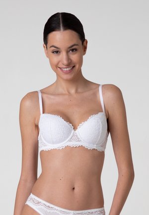 Donna sorridente che indossa un reggiseno in pizzo bianco e biancheria intima abbinata su uno sfondo grigio uniforme.