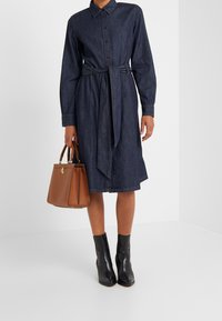 Jeanskjolklänning med bälte i midjan, långa ärmar och knappstängning. Kombineras med en brun läderhandväska och svarta ankelboots.