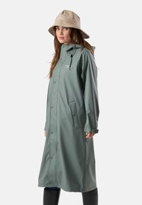 Kvinde iført en lang grå regnfrakke med trykknapper og en beige bucket-hat, stående og kigger let til siden.