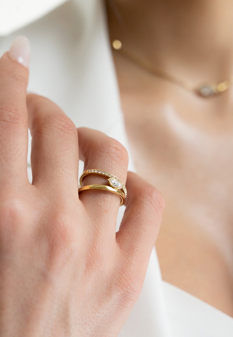 Anelli d'oro con una pietra a goccia e una fascia decorata con piccole pietre trasparenti, indossati da una mano con una manicure rosa chiaro.