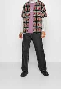 Camicia a maniche corte multicolore con motivi geometrici sopra una maglietta viola, abbinata a pantaloni larghi neri e scarpe chunky nere.
