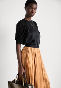 Blouse en satin noir avec des manches bouffantes, associée à une jupe plissée longueur cheville de couleur beige. Sac noir tissé tenu par la main du modèle.