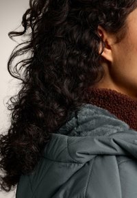 Osoba s tamnom kovrčavom kosom koja nosi plavo-sivu prošivenu jaknu s mekim svijetloplavim podstavom i smeđi, pahuljasti rol-kragnu.