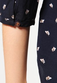 Manica di camicia navy con bottone, caratterizzata da un motivo di foglie chiare sparse; tessuto leggero con una texture morbida.