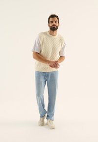 Outfit de bărbați format dintr-un pulover fără mâneci din fir cu model împletit de culoare crem, purtat peste o cămașă albă, asociat cu blugi de culoare albastru deschis și adidași bej.