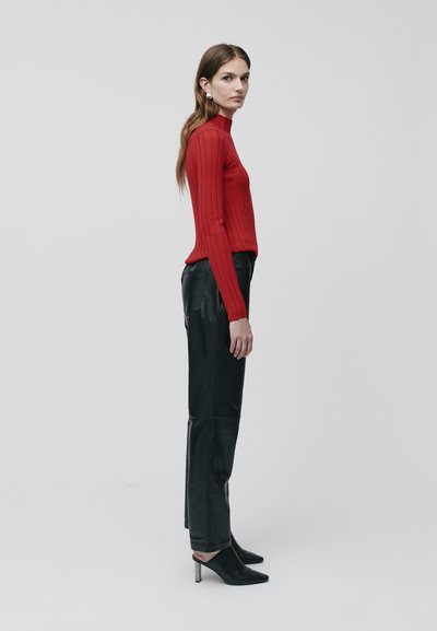 Rdeča rebrasta turtleneck jopica v kombinaciji s črnimi usnjenimi hlačami. Manekenka nosi koničaste črne mule z metalnim peto. Ravninska stranska profilna slika.