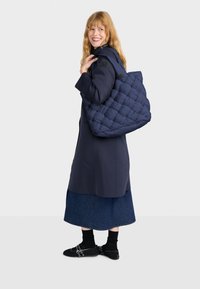 Marineblauwe gewatteerde tote bag met een ruime indeling en handvatten aan de bovenkant, gedragen op de schouder. Model in een lange jas en denimrok, glimlachend.