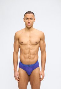 Μυώδης άνδρας με κοντά κούρεμα, φοράει μοβ εσώρουχα Calvin Klein, στέκεται μπροστά σε απλό λευκό φόντο.