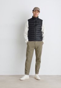 Svart pufferväst över en vit långärmad tröja, matchad med olivgröna byxor och vita sneakers. Modellen bär en beige keps.