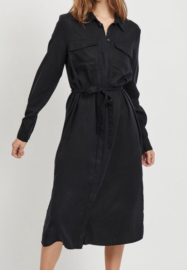 Femme portant une robe chemise midi noire à manches longues avec boutons sur le devant, poches poitrine, et ceinture nouée à la taille, debout devant un fond uni.