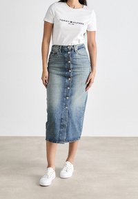 Jeanskjol med hög midja, knäppning framtill och knälång skärning; tillsammans med en vit t-shirt med logotyp och vita sneakers.