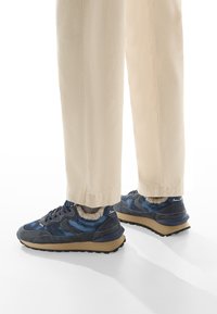Sneakers blu navy con tomaia in materiali misti, dotati di fodera in morbido peluche, suola marrone chiaro e accenti neri. Visibili pantaloni tapered beige.