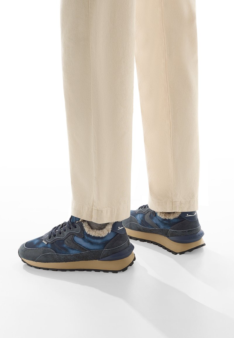 Sneakers blu navy con tomaia in materiali misti, dotati di fodera in morbido peluche, suola marrone chiaro e accenti neri. Visibili pantaloni tapered beige.