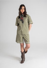 Kaki skjorte med korte ermer og matchende shorts med blomstermønster på kanten. Modell iført brune cowboy-støvler og et mønstrete halsbånd.