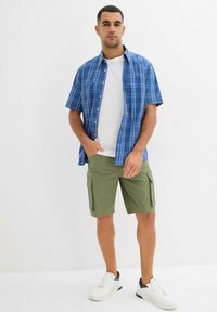 Camicetta a maniche corte a scacchi blu sopra una t-shirt bianca, abbinata a pantaloni cargo verde oliva e sneakers bianche con dettagli neri.