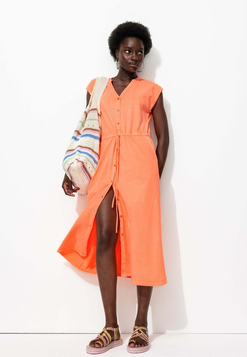 Robe chemise corail orangé avec taille réglable, fente jusqu'aux genoux et manches courtes. Associée à un sac en tricot coloré et des sandales.