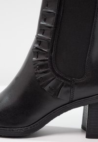 Gros plan d'une bottine noire en cuir avec panneau latéral élastique, bordure froncée décorative et talon bloc.