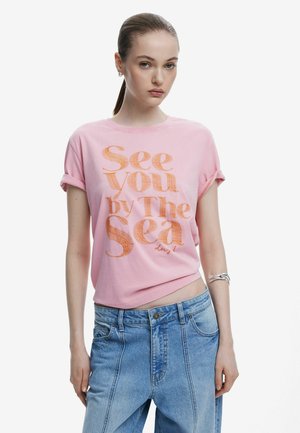 T-shirt en coton rose avec des manches retroussées, arborant le texte "See You by The Sea" en orange. Associé à un jean en denim léger bleu clair taille haute.