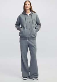 Sudadera gris con capucha, botones en la parte delantera y bolsillos, combinada con pantalones de pierna ancha a juego. La tela parece suave y texturizada. Detalles en negro.