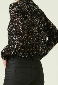 Blusa a fantasia bianco e nero con una texture trasparente, dotata di polsini con bottoni e retro arricciato, abbinata a jeans neri aderenti.
