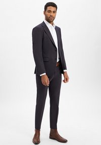 Marineblaues Sakko mit zwei Knöpfen, passender Hose, weißem Hemd und braunen Lederstiefeln bis zum Knöchel. Glattes Material, maßgeschneiderte Passform.
