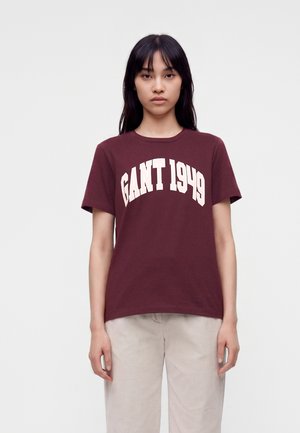 Femme aux longs cheveux foncés portant un T-shirt bordeaux avec "GANT 1949" en texte blanc et un pantalon clair, debout devant un fond uni.