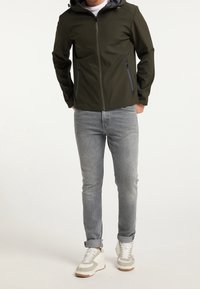 Olivgrön softshell-jacka med huva och dragkedjefickor, i kombination med ljusgrå jeans och vita sneakers med beige accenter.