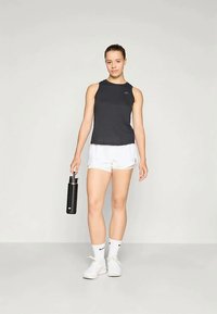 Schwarzes ärmelloses Sportoberteil, weiße Shorts, hält eine schwarze Wasserflasche. Weiße Sneakers, Knöchelsocken. Einfaches Design mit minimalen Details.