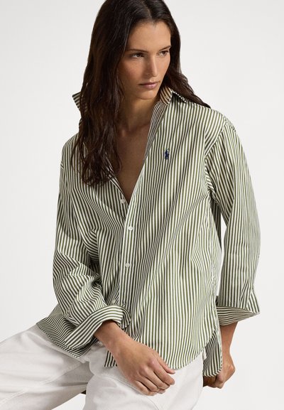 Γυναίκα με μακριά σκούρα μαλλιά, ντυμένη με φαρδιά μπλούζα με ριγέ πράσινο και λευκό και λευκό παντελόνι, καθισμένη μπροστά σε απλό φόντο.