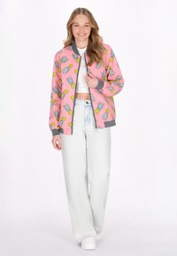 Jachetă bomber roz cu un model de ananas și frunze de palmier, cu manșete și tiv gri culcat, purtată peste un top scurt alb și blugi deschiși.