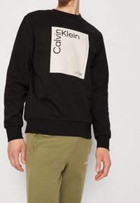 Felpa nera con un grande logo rettangolare bianco che presenta "Calvin Klein" e "Est. 1968" in carattere grassetto. Tessuto morbido, scollatura a girocollo.