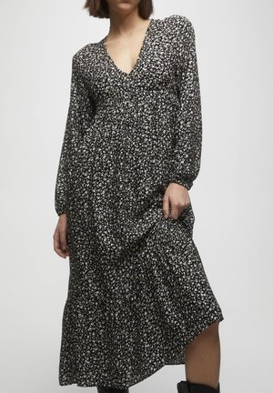 Femme portant une robe midi noire à manches longues avec un imprimé floral blanc, tenant légèrement relevée la jupe sur un côté.