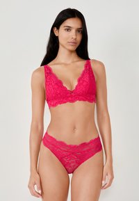 Różowy koronkowy biustonosz typu bralette z głębokim dekoltem w serek i falistymi brzegami, w komplecie z pasującymi różowymi koronkowymi majteczkami hipster, ozdobionymi złożonymi wzorami.
