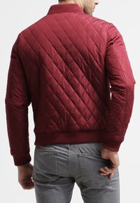 Burgundy quilted bomber jacket with a ribbed collar and cuffs. Features diamond patterns and a fitted waist. Made from shiny, smooth fabric.